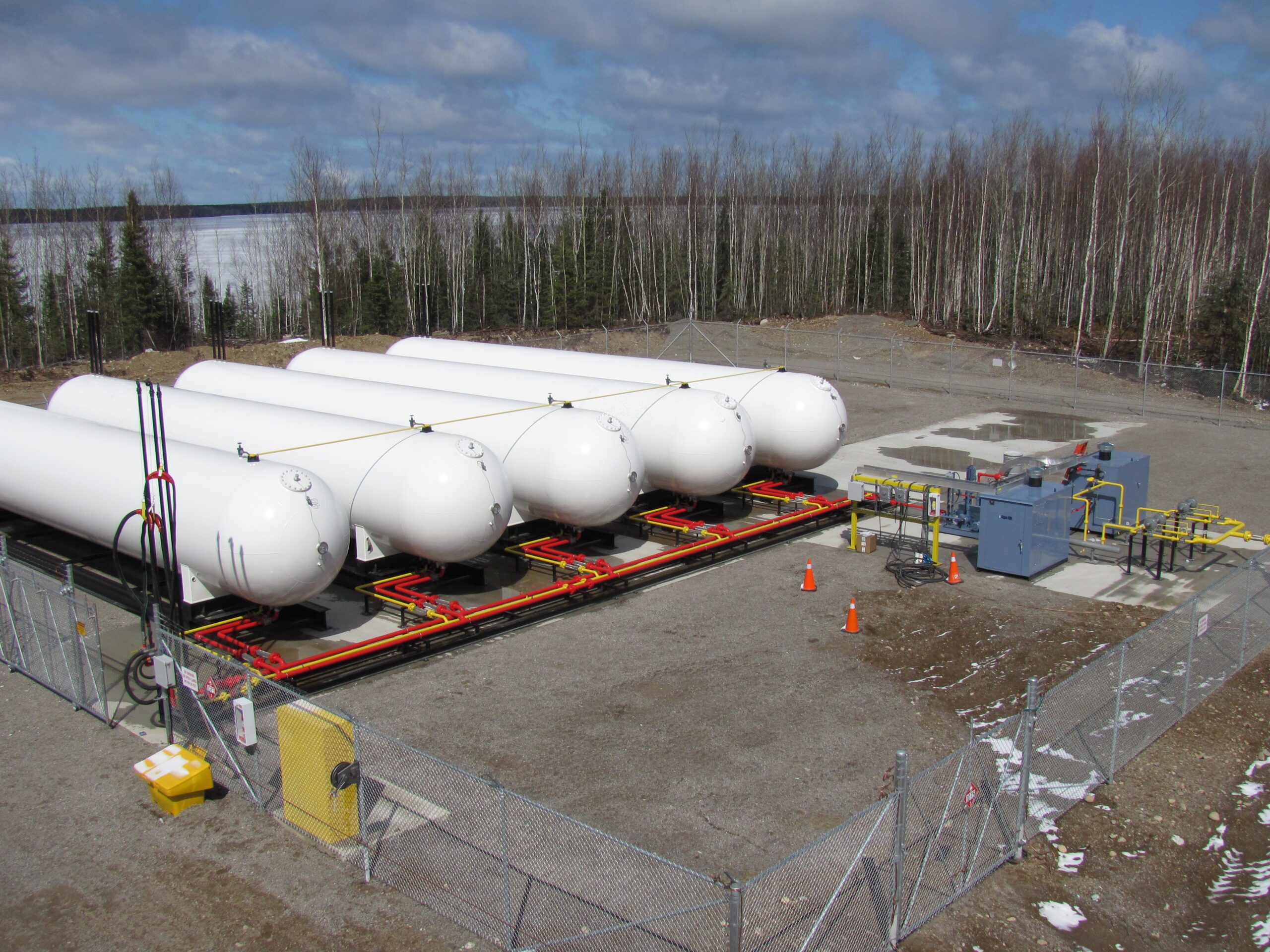 Several propane tanks arranged in a row on a gravel surface. The tanks are large and cylindrical in shape, with a silver metal exterior and black caps. Some tanks are partially covered with yellow caution tape. In the background, there are trees and bushes visible